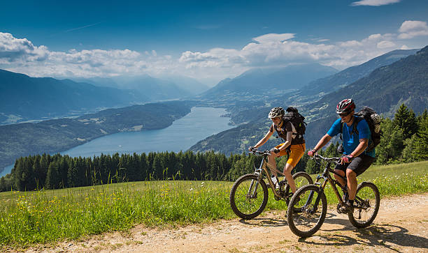 A couple is biking uphill on a scenic forest road with an overview of Lake Millstatt, Carinthia, southern Austria. The region is a very attractive holiday area and is famous for hiking, mountainbiking and swimming during summertime. In wintertime there lots of skiing areas as well.