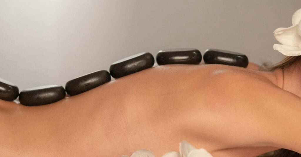 massage-bien-etre Une femme est allongée avec des pierres médicinales sur le dos et des fleurs de thiaré pour un massage.