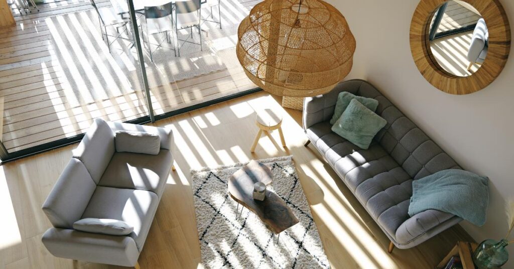 Un salon cosy avec deux canapés, une table basse, un tapis, une suspension boule en rotin, un grand miroir rond en bois et de larges baies vitrées. 