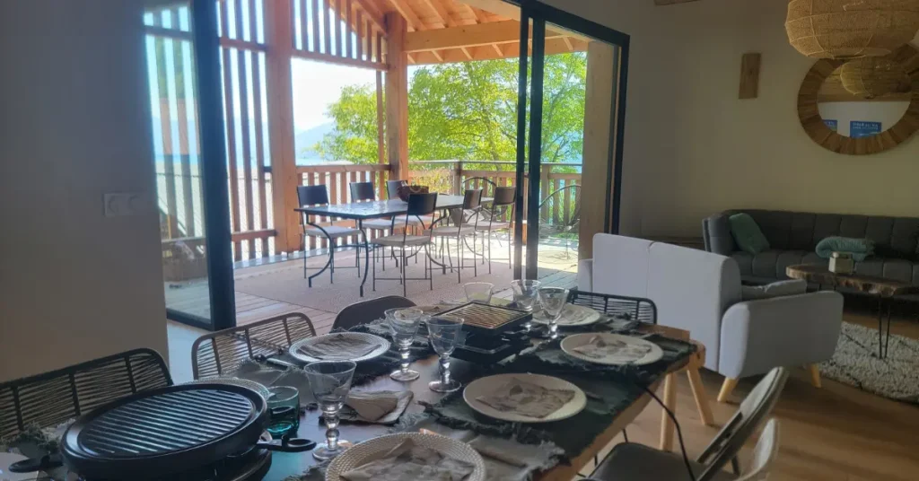 Une table est dressée devant une large baie vitrée ouverte sur une terrasse en bois avec vue lac. La table attend les participants à un séminaire au vert chez Les Granges de Capucine.